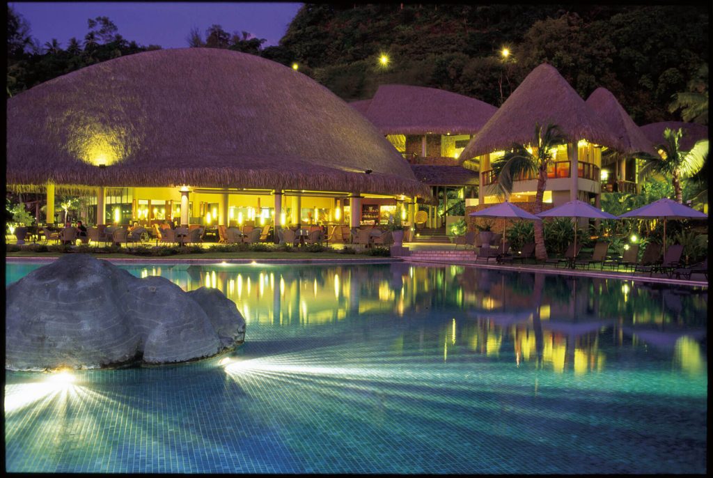 Nighttime Poolside at Le Tahiti by Pearl Resorts, Featuring Illuminated Infinity Pool, Tropical Landscaping, and Views of Matavai Bay Under a Starlit Sky.