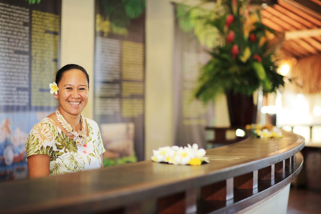 | Blue Tahiti Travel Warm Welcome at the Reception Desk of Le Tahiti by Pearl Resorts, Where Friendly Staff Greet Guests with Traditional Polynesian Hospitality in a Tropical Setting.