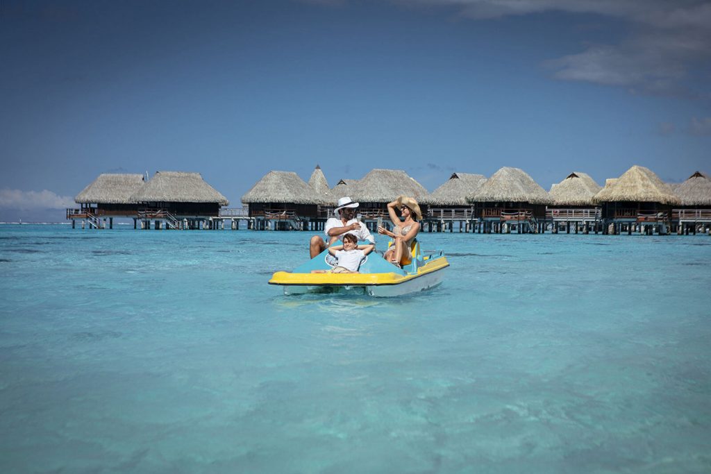 | Blue Tahiti Travel Australian Family Kayaking Together in the Clear Lagoon Waters Near Sofitel Kia Ora Moorea Beach Resort, with Overwater Bungalows in the Background.