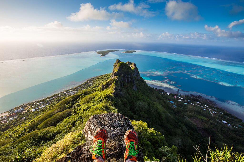 | Blue Tahiti Travel Hiker Ascending the Ridge Trail of Mount Teurafaatiu on Maupiti Island, with Panoramic Views of the Turquoise Lagoon and Surrounding Ocean.